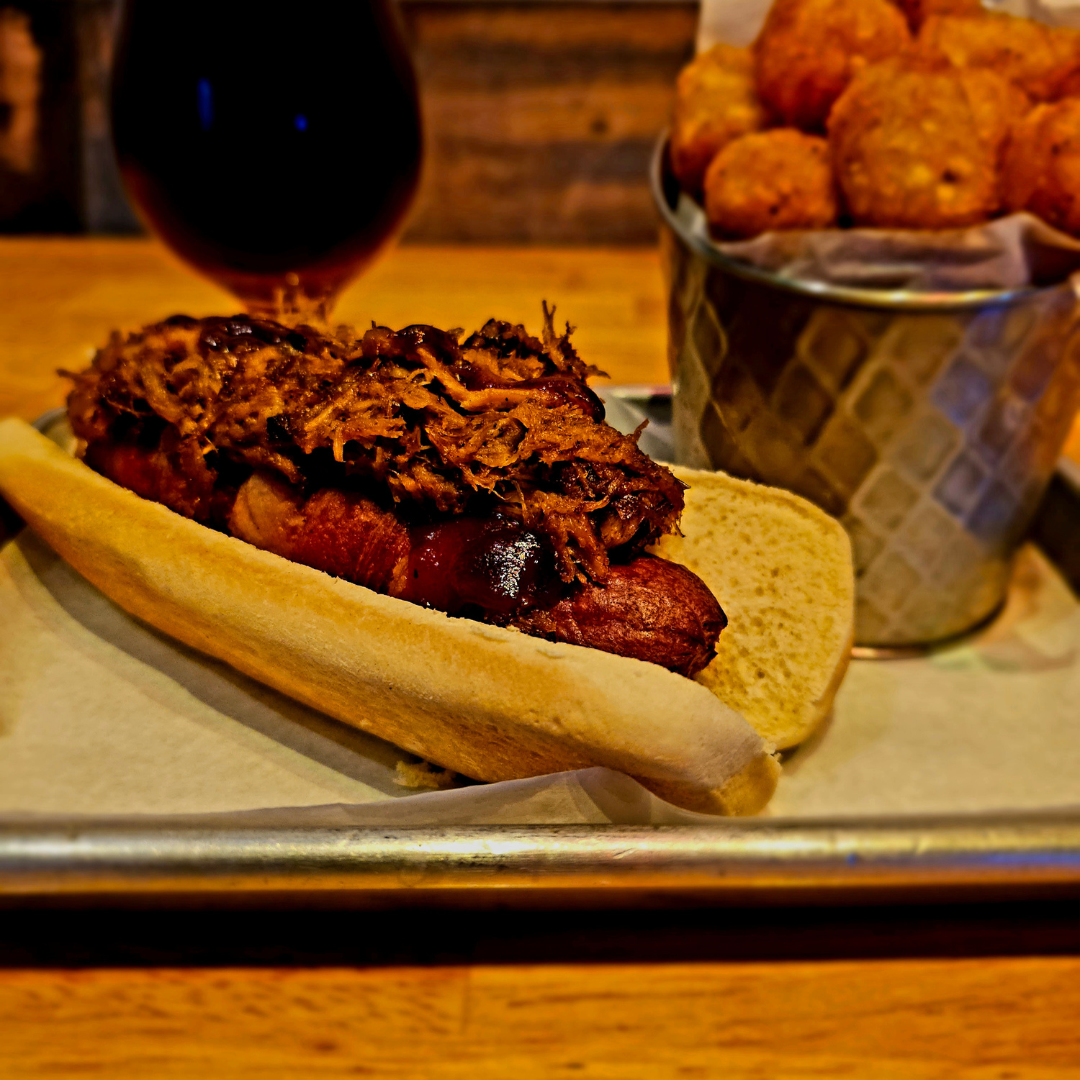 A close-up of a mouthwatering Fort Worth hot dog at Bowlounge, featuring a bacon-wrapped glamour dog topped with savory pulled pork and a generous drizzle of BBQ sauce, accompanied by a side of crispy fries or tots.