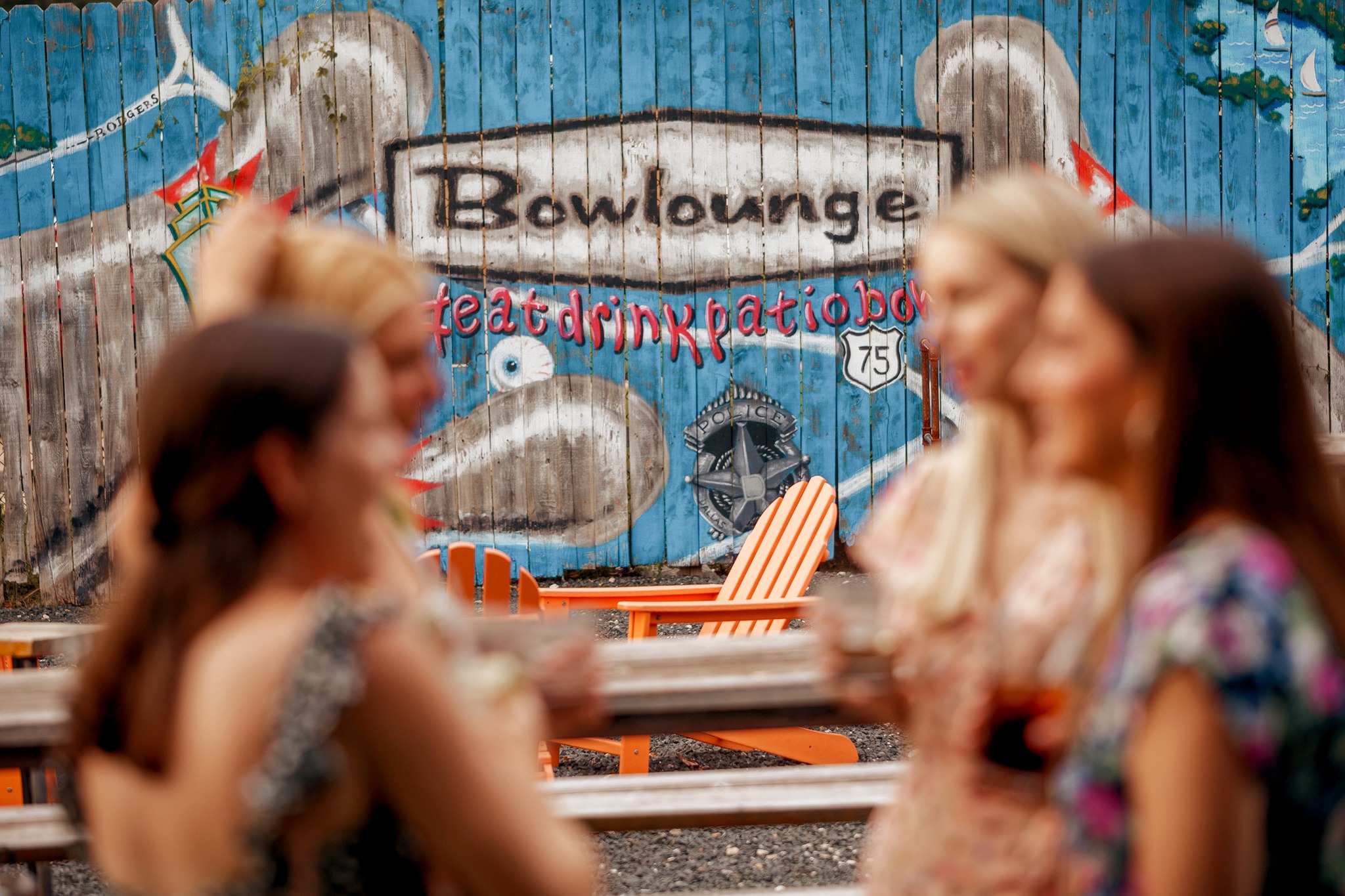 women engaged in conversation in front of Bowlounge Dallas's vibrant mural, featuring the logo and bowling pins, setting the stage for a fun-filled outing with friends, delicious drinks, and exciting games.